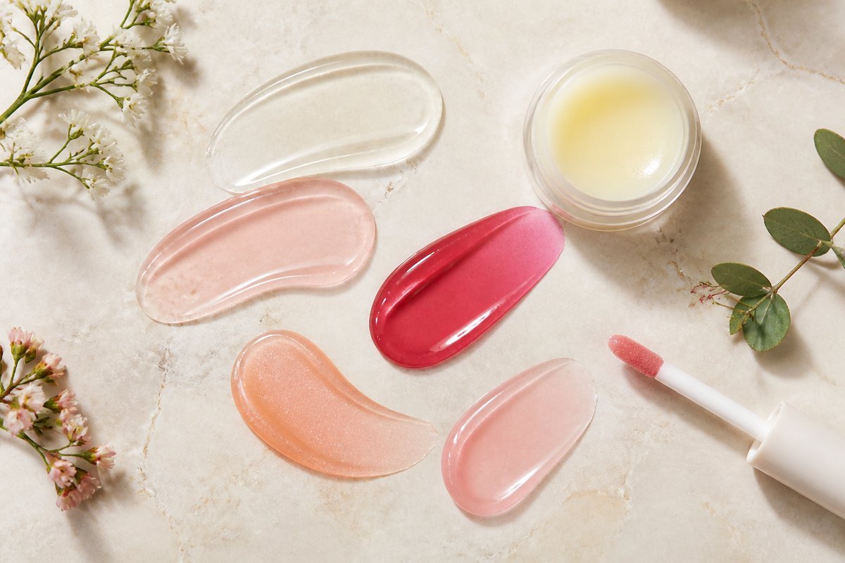 Assorted lip gloss swatches and white gloss pot on marble surface with flowers